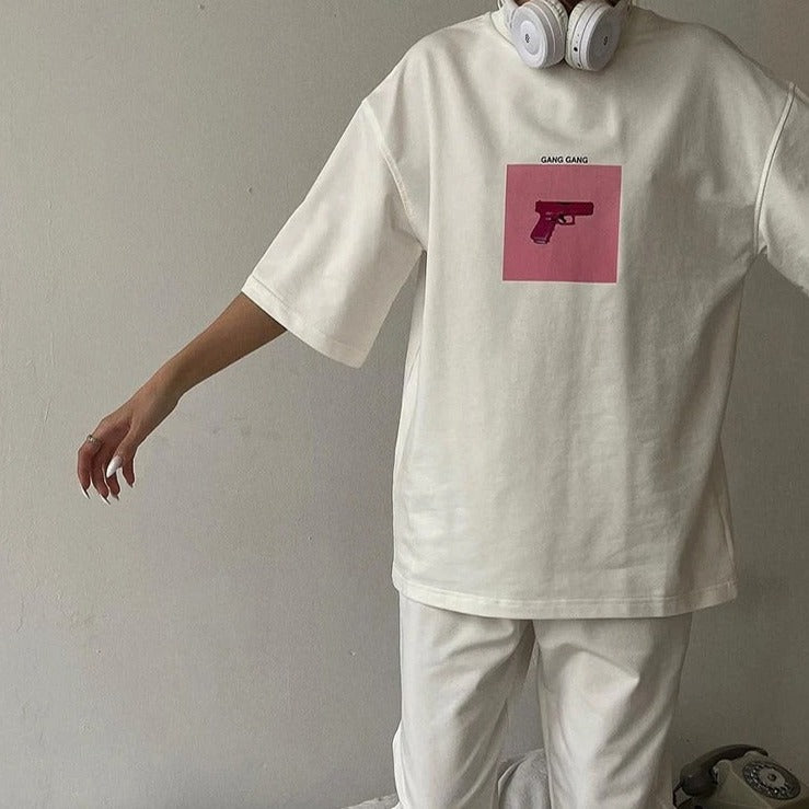 Weißes Oversize-T-Shirt mit rosa Pistolen-Print, kombiniert mit weißen Kopfhörern. Trendige Streetwear-Mode für Damen.
