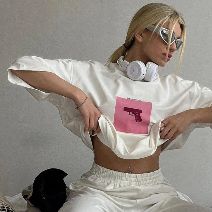 Frau in weißem Outfit mit Kopfhörern und Sonnenbrille, zeigt ein T-Shirt mit pinkem Pistolen-Design. Trendige Mode, urbaner Stil, Streetwear.