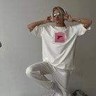 Frau in weißem Oversize-T-Shirt mit pinkem Print, Sonnenbrille und Kopfhörern. Trendige Streetwear, lässiger Look, modisches Statement.