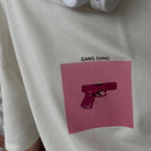 Graues T-Shirt mit pinkem Pistolen-Print und "Gang Gang"-Schriftzug. Trendige Streetwear, urbaner Stil, modisches Design.