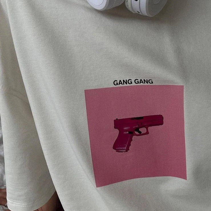 Graues T-Shirt mit pinkem Pistolen-Print und "Gang Gang"-Schriftzug. Trendige Streetwear, urbaner Stil, modisches Design.