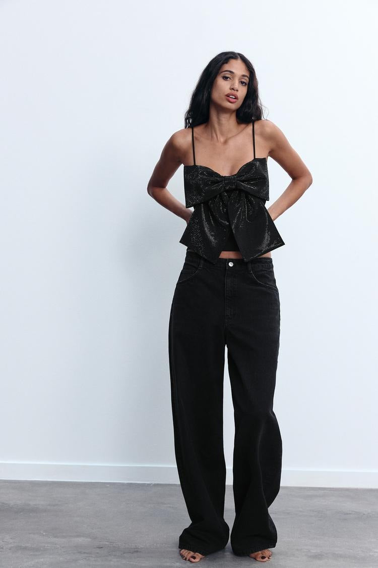 Frau in schwarzem, glitzerndem Top mit Schleife und weiten Jeans. Modetrend, eleganter Look, Damenmode, stilvolle Outfits, Freizeitkleidung.