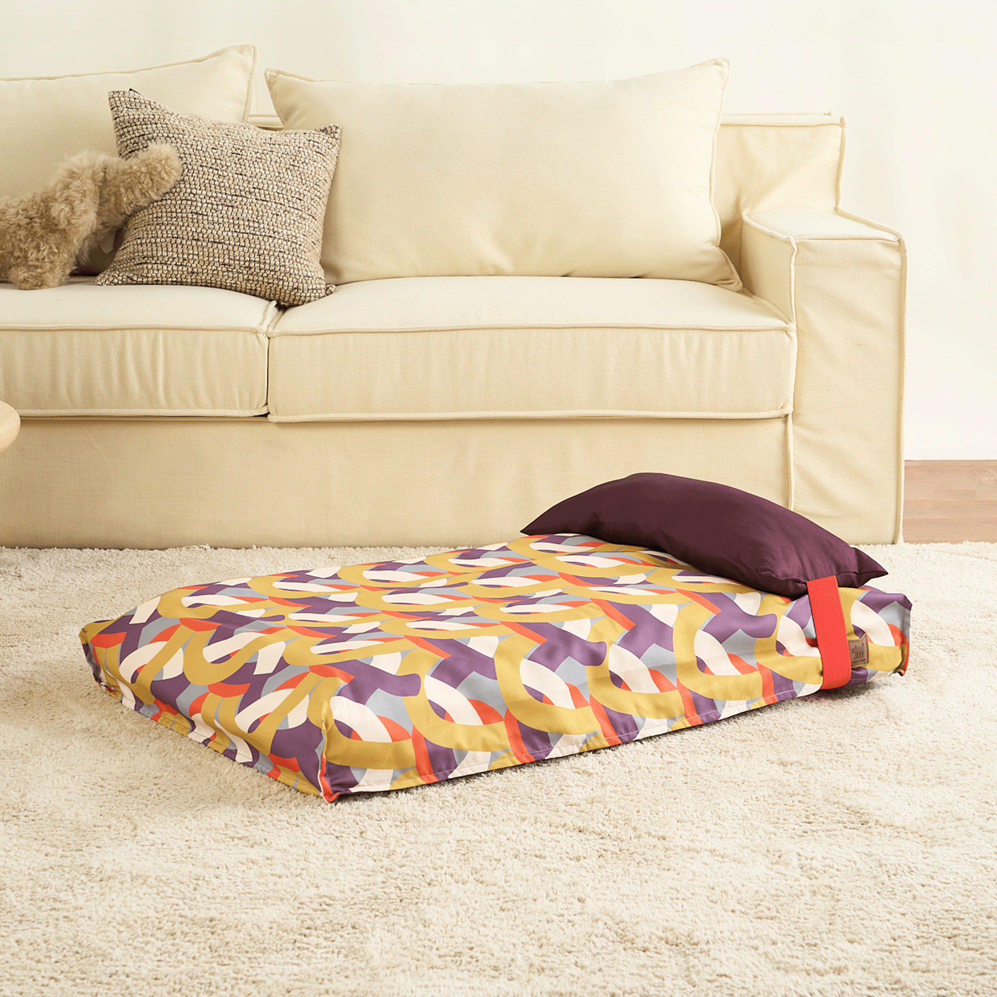 Buntes Hundebett mit geometrischem Muster, Kissen, auf beigem Teppich vor Sofa.