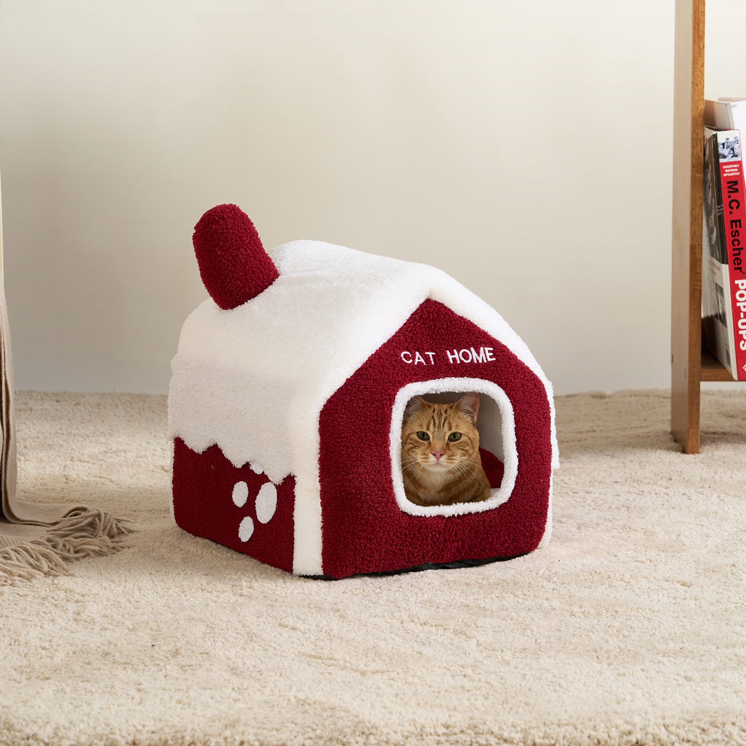 Rotes Katzenhaus aus Plüsch, schneebedeckt, mit Katze im Inneren, auf Teppichboden.