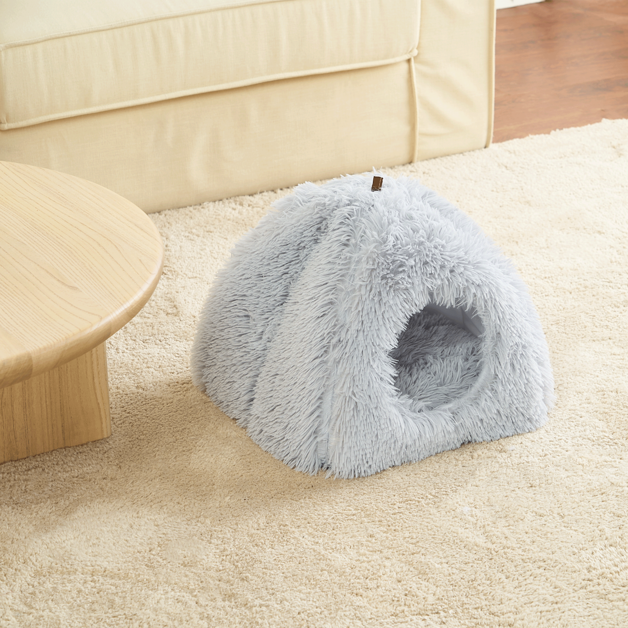 Graue flauschige Haustierhöhle auf beigem Teppich neben Holztisch im Wohnzimmer.