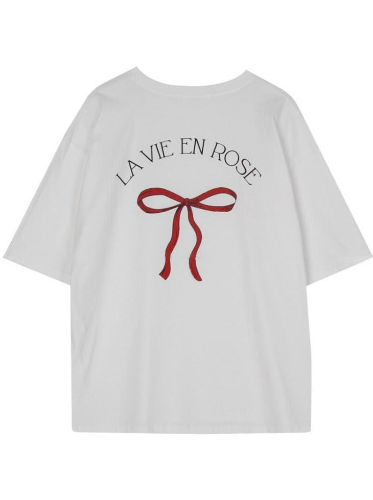 Weißes T-Shirt mit roter Schleife und "La Vie en Rose" Aufdruck. Modisches Damenoberteil, ideal für lässige Outfits. Hochwertige Baumwolle.