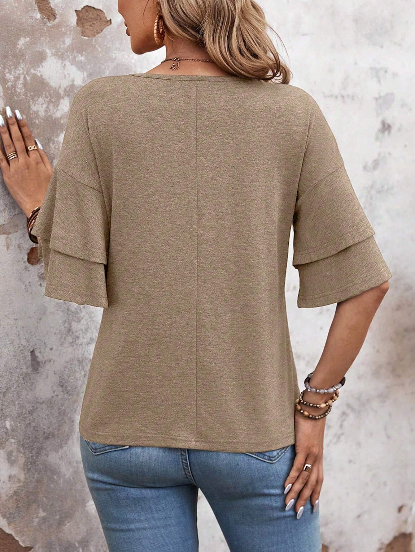 Frau in beige Bluse mit Rüschenärmeln und blauer Jeans vor rustikaler Wand.