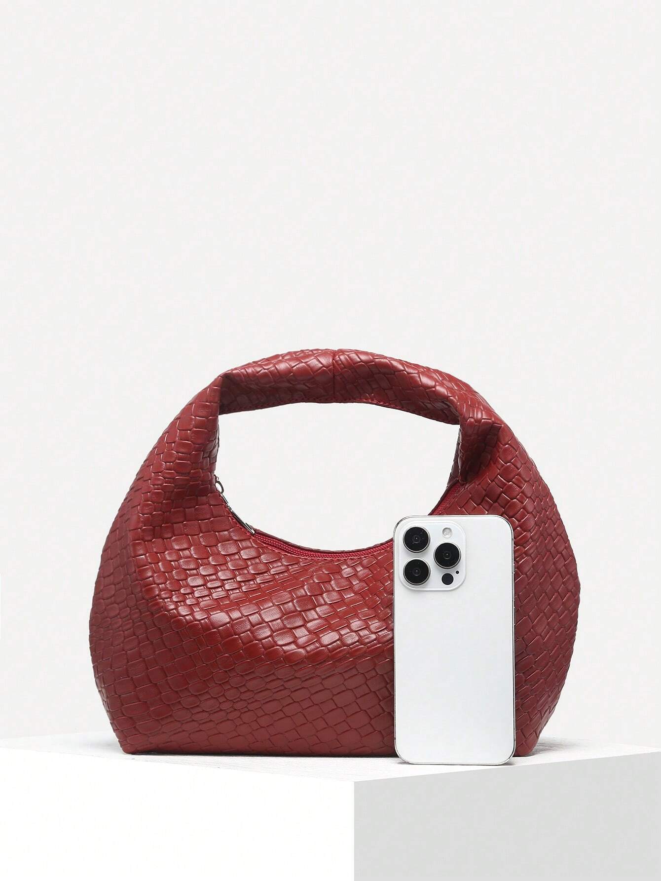Rote geflochtene Handtasche aus Leder neben einem weißen Smartphone auf weißem Hintergrund.
