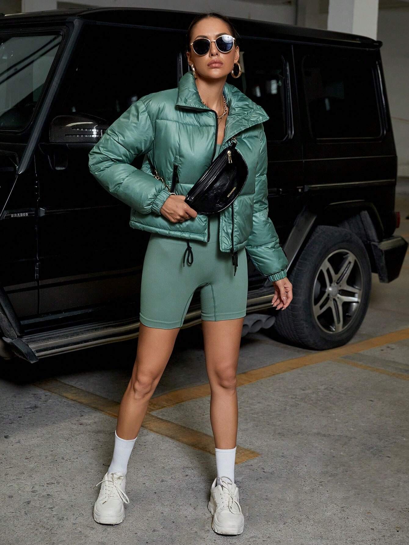 Frau in grüner Steppjacke und Shorts, schwarze Gürteltasche, Sonnenbrille, vor schwarzem SUV.