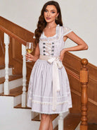 Frau in traditionellem Dirndl mit Spitzenmieder und Schleife, hält ein Glas Weißwein, steht auf Holztreppe. Oktoberfest, Trachtenmode, elegant.