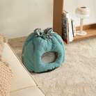 Blaues Katzenbett aus Stoff, gemütlich, rund, auf Teppichboden neben Sofa und Regal.