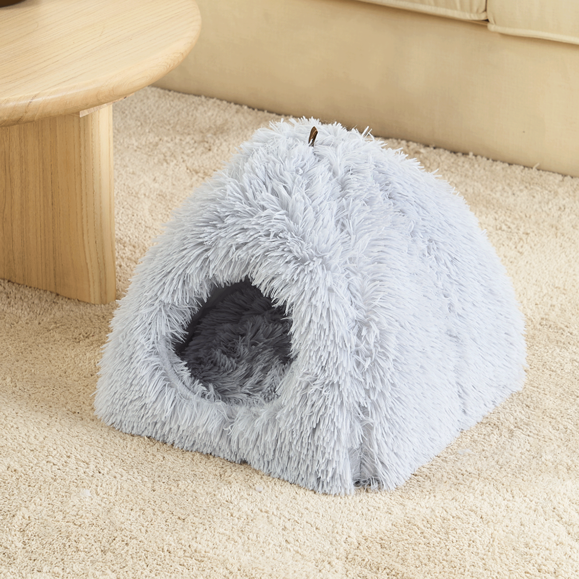 Graues, flauschiges Haustierbett in Höhlenform auf beigem Teppich neben Holztisch.