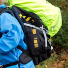 Person mit blauem Anorak trägt schwarzen und neongelben Wanderrucksack im Wald.