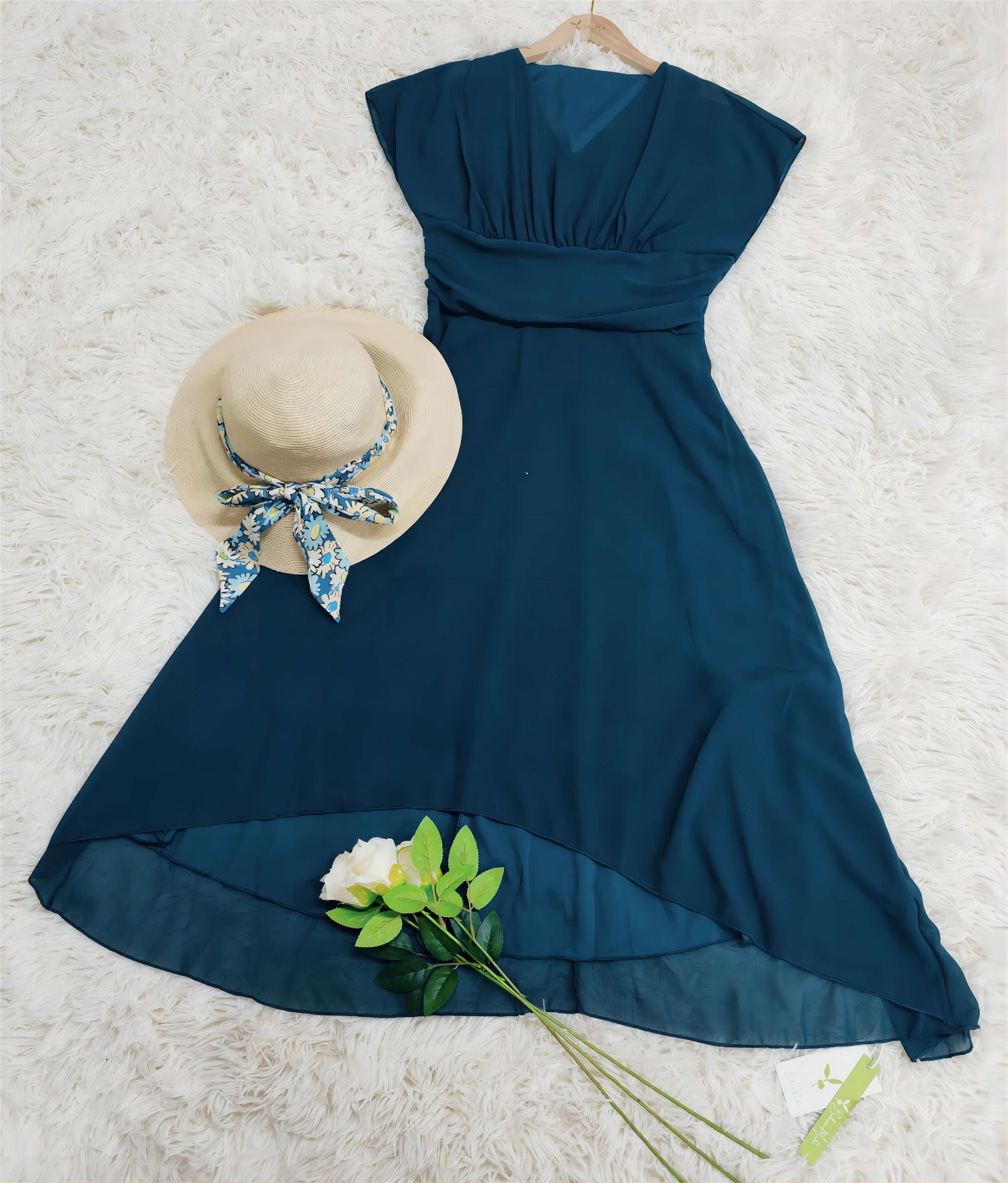 Elegantes blaues Kleid mit asymmetrischem Saum auf weißem Teppich, kombiniert mit Strohhut und Blumen. Perfekt für Sommermode und Damenbekleidung.