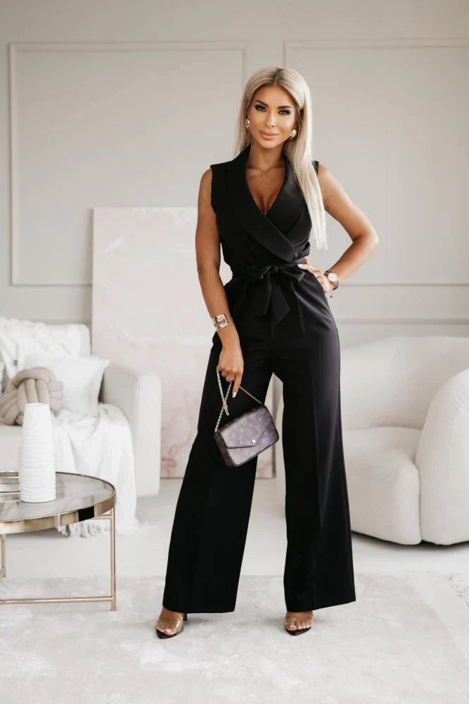 Frau in schwarzem ärmellosen Jumpsuit, hält eine kleine Handtasche, steht in modernem Wohnzimmer.