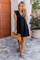 Frau in schwarzem Sommerkleid mit Strohtasche, steht vor Holztür und blühenden Pflanzen. Modetrend, Sommeroutfit, Damenmode, eleganter Look.