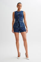 Frau in ärmellosem Jeans-Set mit Knöpfen, bestehend aus Weste und Shorts, kombiniert mit weißen High Heels. Trendige Damenmode, eleganter Look.