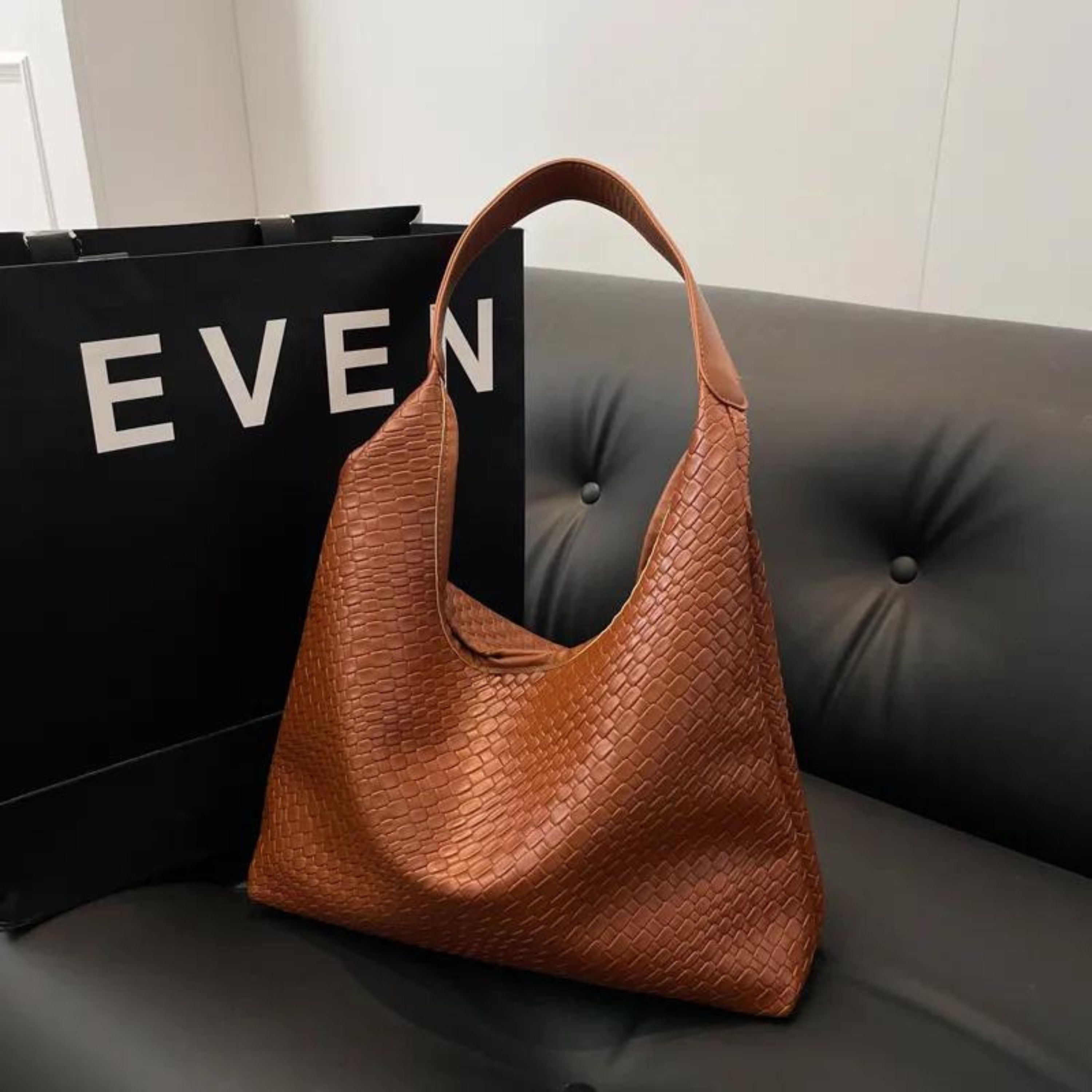 Braune Lederhandtasche mit geflochtenem Design auf schwarzem Sofa, neben einer Einkaufstasche. Stilvoll, elegant, modisch, Damenaccessoire.