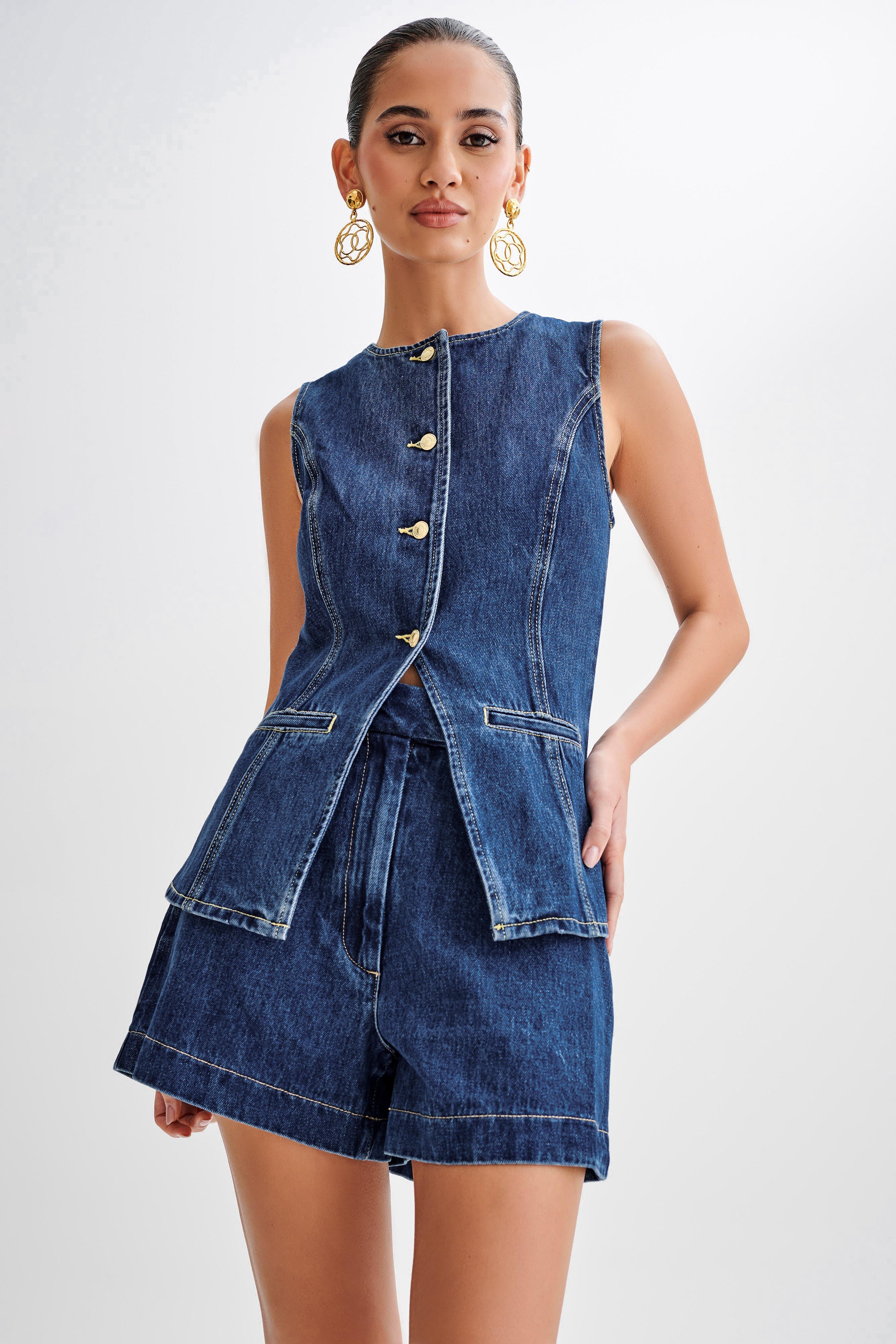 Frau in ärmelloser Jeansjacke und Shorts, goldene Knöpfe, elegante Ohrringe. Modetrend, Damenbekleidung, lässiger Chic, Denim-Outfit.