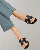 Schwarze Ledersandalen mit markantem H-Design, getragen von einer Person in blauer Hose. Modische Sommerschuhe, ideal für stilvolle Outfits.