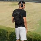 Mann mit schwarzem Rucksack und Kopfhörern im Freien, trägt schwarzes T-Shirt und weiße Shorts.