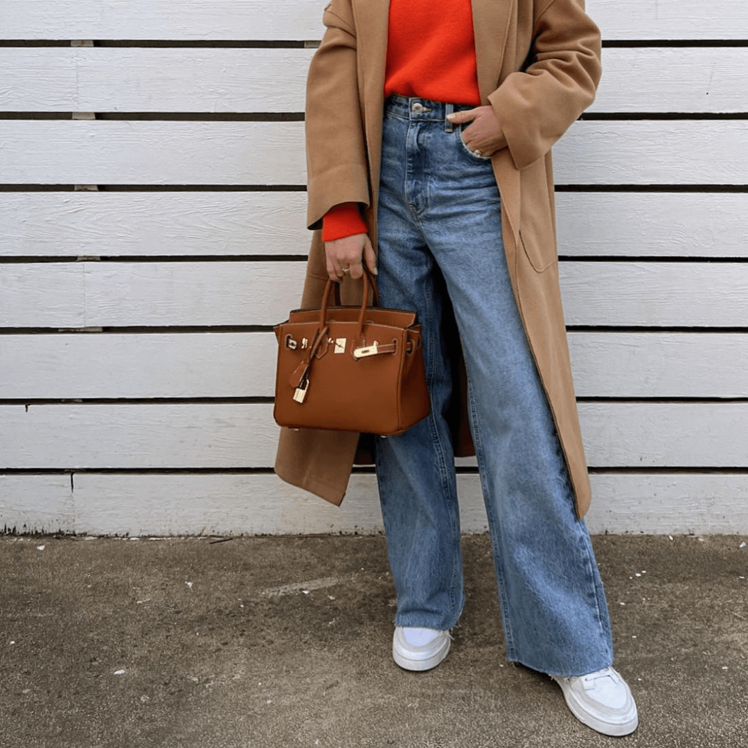 Frau mit braunem Mantel, rotem Pullover, blauer Jeans und brauner Handtasche vor Holzwand.