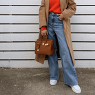 Frau mit braunem Mantel, rotem Pullover, blauer Jeans und brauner Handtasche vor Holzwand.