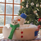 Katzenbett in Schneemannform, beige, mit grünem Schal, neben Weihnachtsbaum, gemütlich, dekorativ.