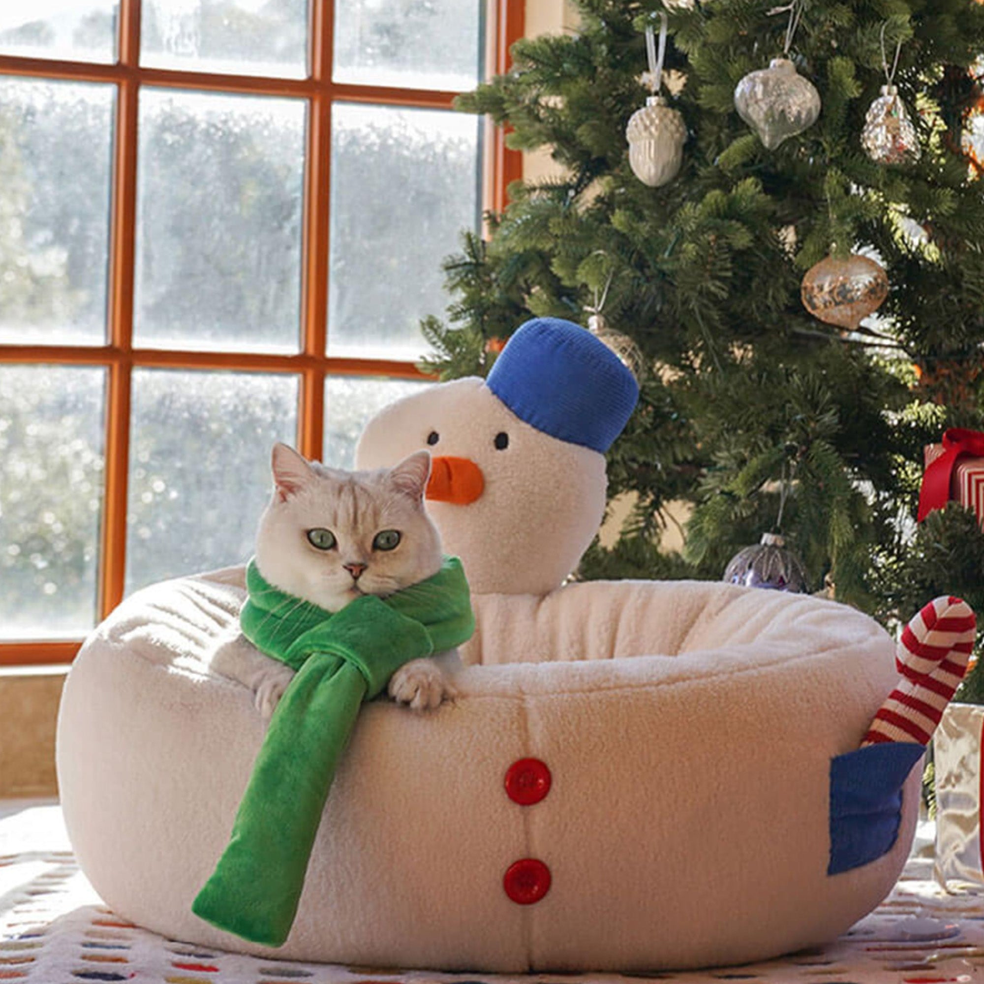 Katzenbett in Schneemannform, beige, mit grünem Schal, neben Weihnachtsbaum, gemütlich, dekorativ.