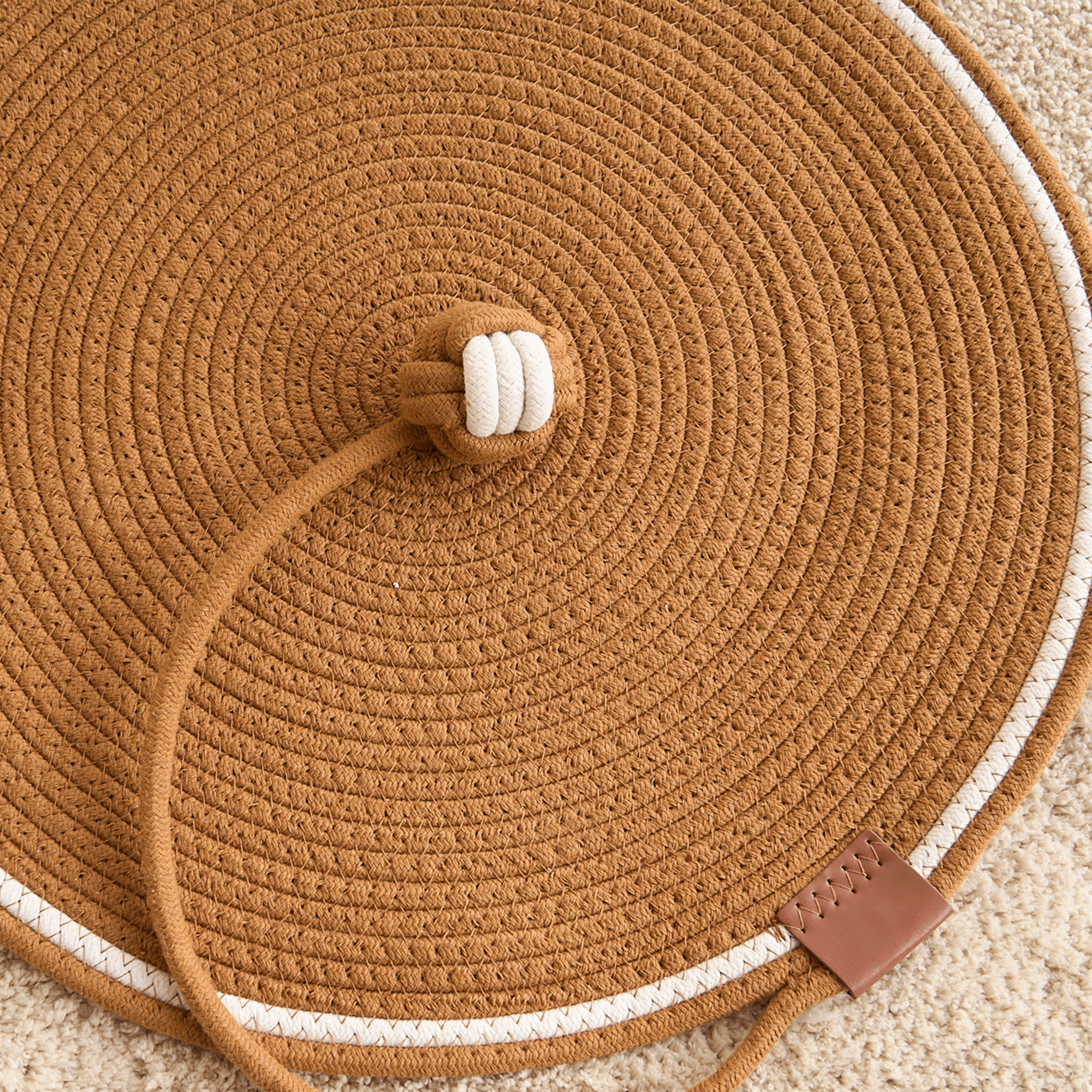 Katzenspielzeug Kratzmatte aus braunem Seil, rund, mit weißem Rand und Ledertag.