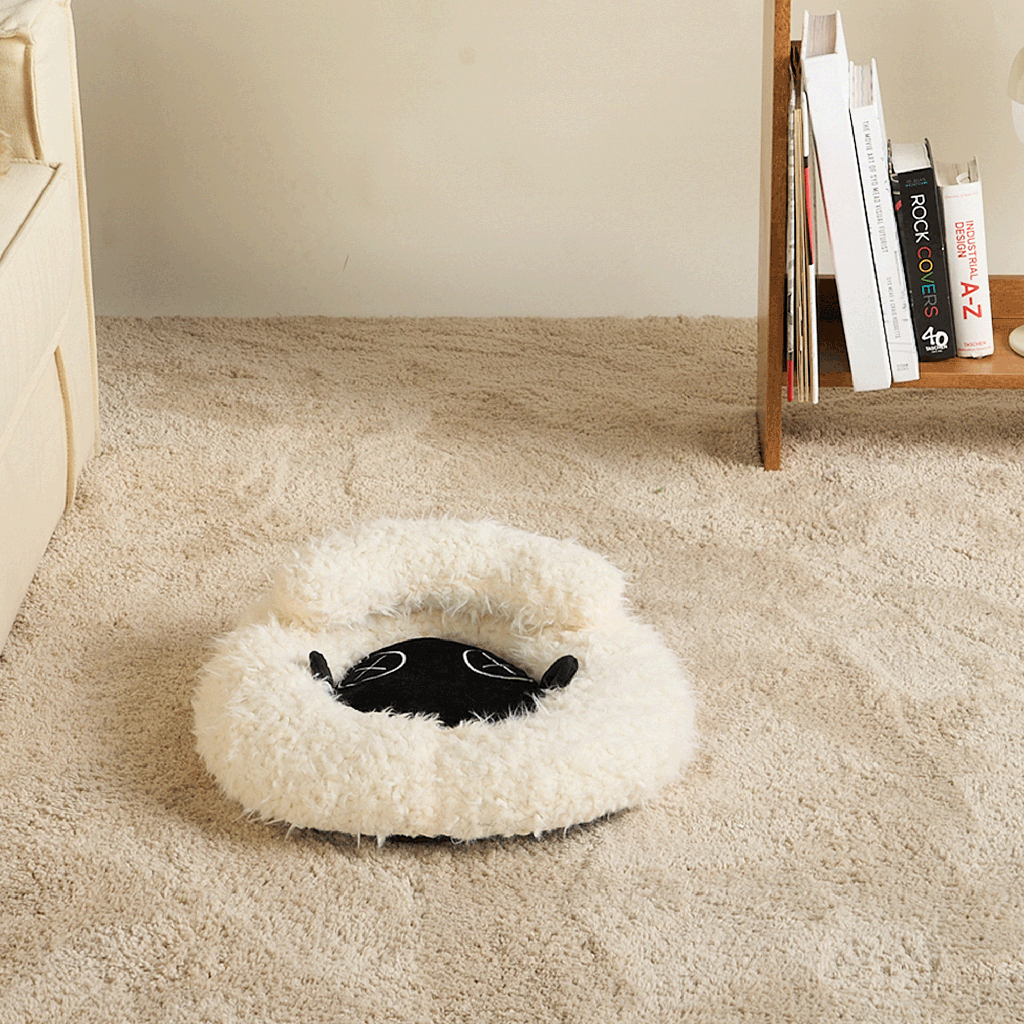 Katzenbett aus weißem Plüsch, rund, flauschig, auf beigem Teppich, neben Bücherregal.