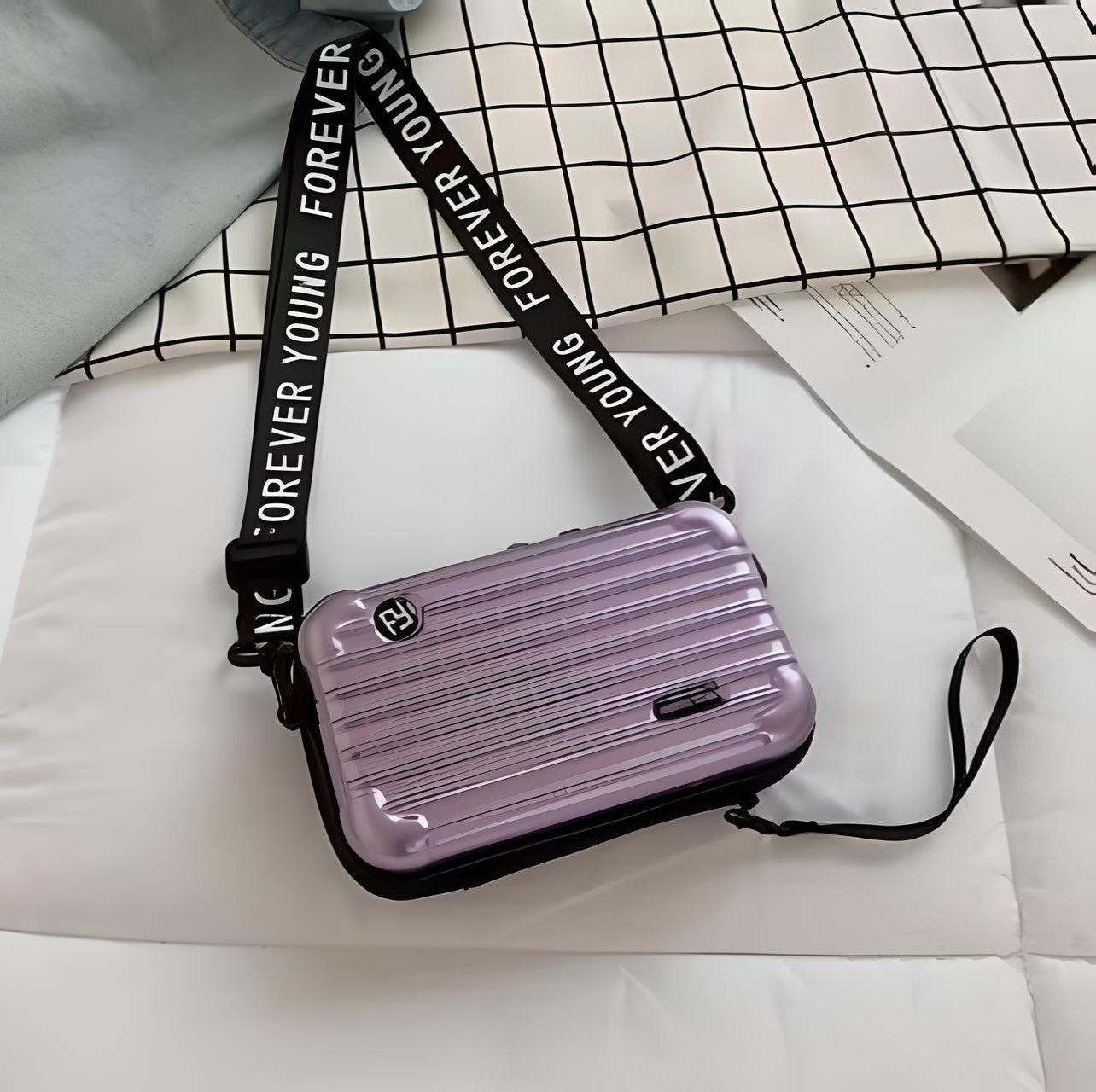 Lila Mini-Koffer Handtasche mit "Forever Young" Gurt auf kariertem Hintergrund.