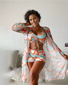 Frau in buntem, gemustertem Strandoutfit mit Kimono und Shorts, lächelnd im Wohnzimmer. Sommermode, Damenbekleidung, Freizeitlook.