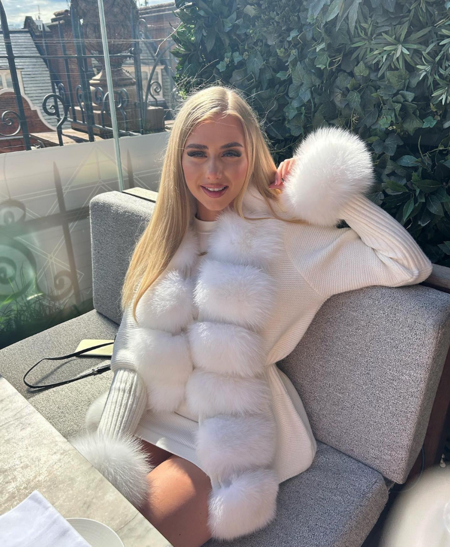 Blonde Frau in weißem, pelzbesetztem Pullover auf Terrasse. Elegante Mode, Herbstoutfit, luxuriöser Stil, Outdoor-Mode, trendige Damenbekleidung.