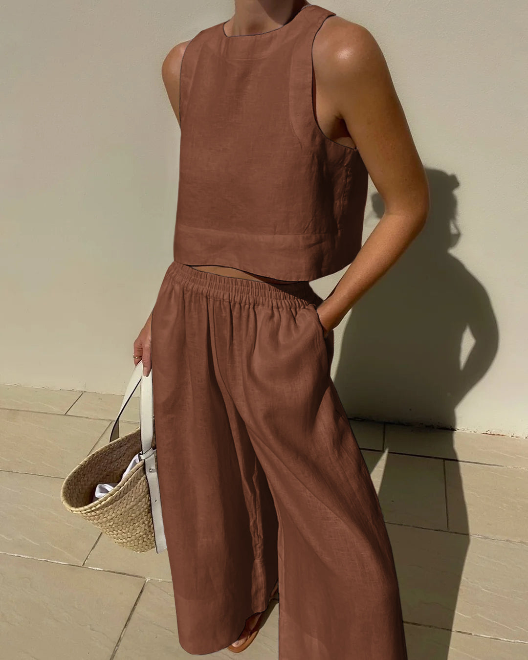 Frau in braunem Leinen-Zweiteiler, bestehend aus ärmellosem Top und weiten Hosen, hält eine Strohtasche. Sommermode, lässiger Stil, Leinenkleidung.