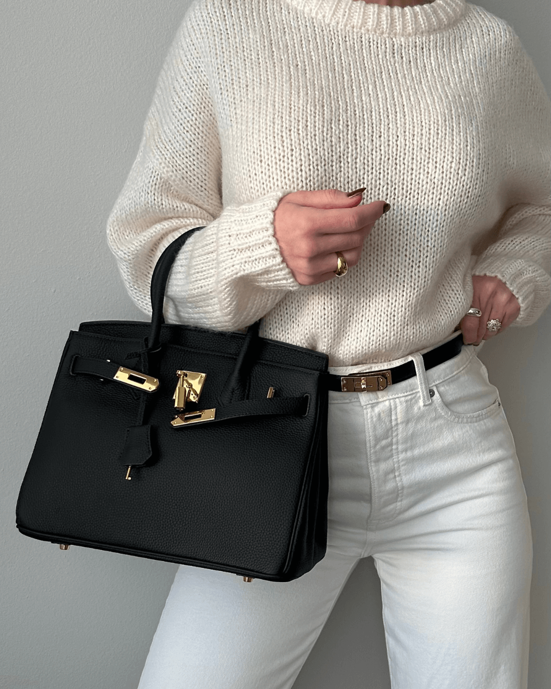 Frau mit schwarzer Lederhandtasche, goldene Details, weißer Pullover, modisch, elegant.
