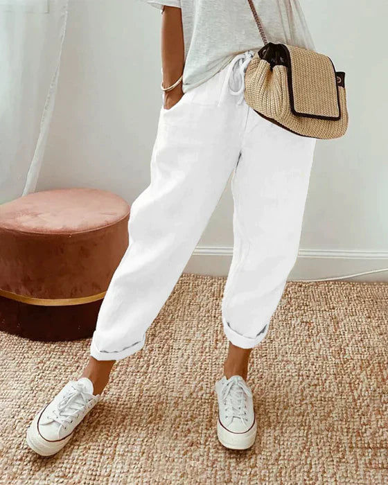 Frau in weißen Leinenhosen, grauem T-Shirt, Strohtasche, weiße Sneaker, modisch, lässig.