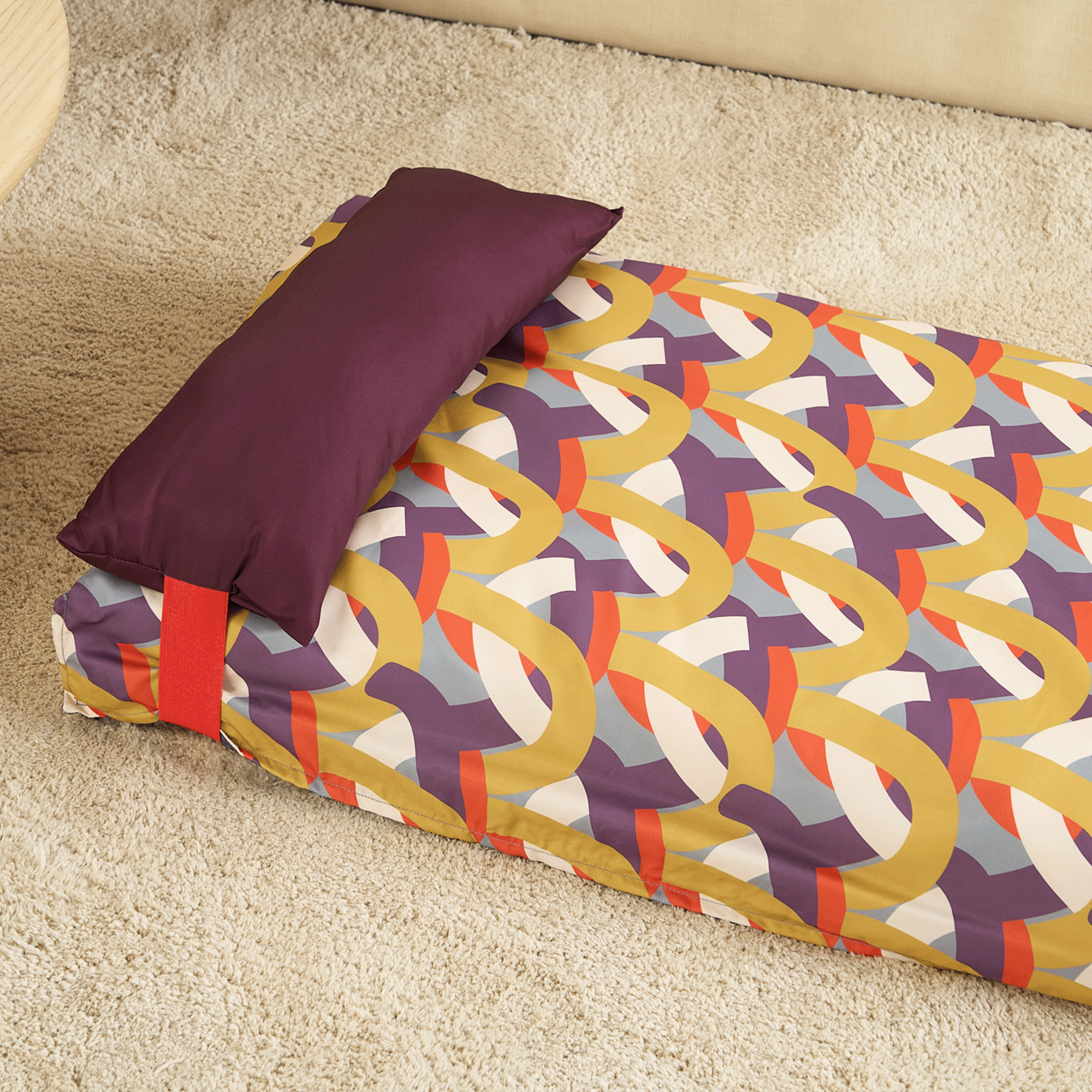 Buntes Hundebett mit geometrischem Muster und lila Kissen auf beigem Teppich.