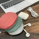 Laptop mit rosa und grüner faltbarer Silikonschüssel, AirPods und Ladekabel auf Holztisch.