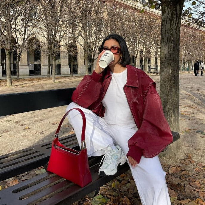 Frau in roter Lederjacke und weißem Outfit sitzt auf Parkbank, trinkt Kaffee. Modetrend, Herbstmode, Streetstyle, rote Handtasche, Sonnenbrille.