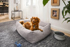 Braunes Hundebett aus Stoff auf grauem Teppich in modernem Wohnzimmer mit Hund und Spielzeug.