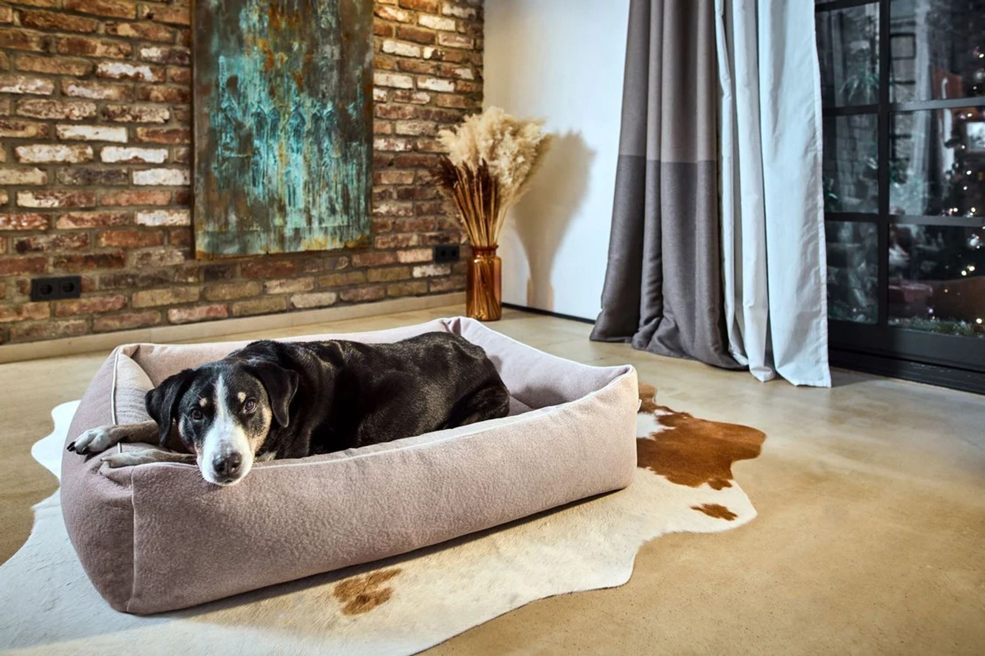 Schwarzer Hund auf grauem Hundebett in modernem Wohnzimmer mit Backsteinwand und Kunstwerk.
