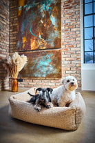 Zwei Hunde auf beigem Hundebett vor Backsteinwand mit Kunstwerk und Trockenblumen.