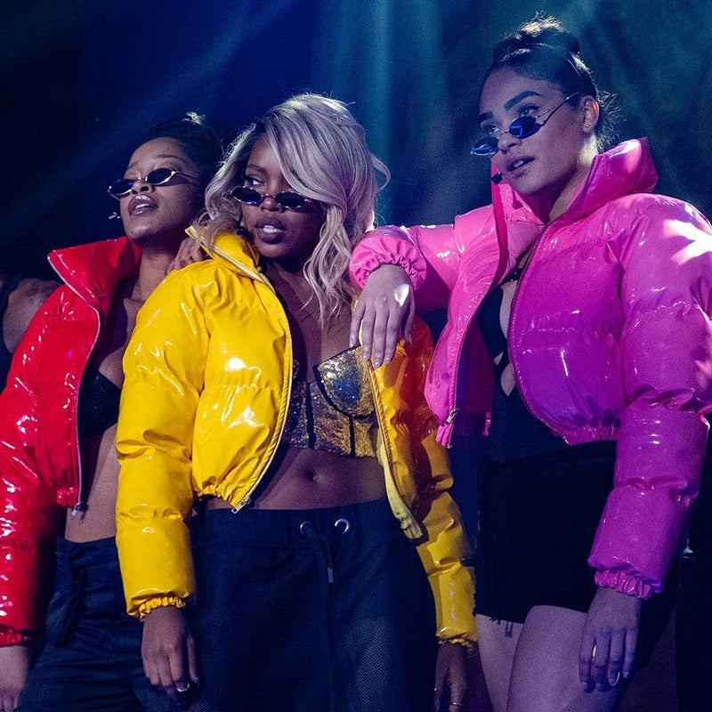 Drei Frauen in glänzenden, bunten Daunenjacken (gelb, pink, rot) und Sonnenbrillen posieren selbstbewusst auf einer Bühne. Fashion, Streetwear, Trend.