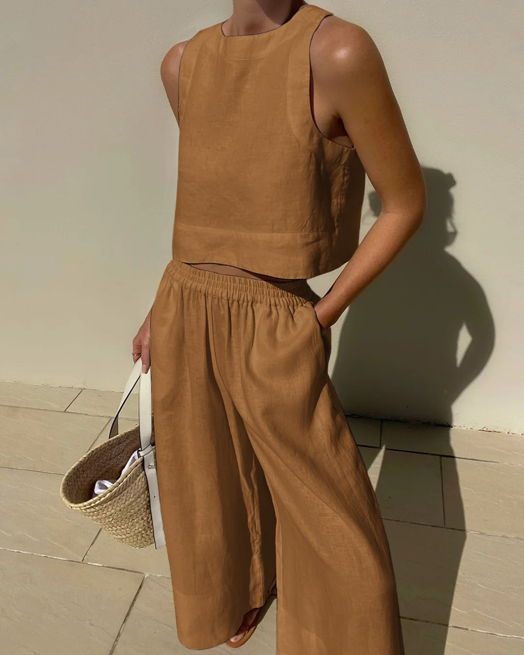 Frau in braunem Leinen-Zweiteiler mit weitem Hosenrock und ärmellosem Top, hält eine Strohtasche. Sommermode, Leinenkleidung, Damenoutfit.