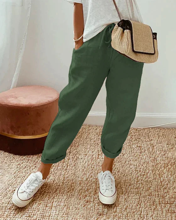 Frau in grüner Leinenhose, weißem T-Shirt, Strohtasche und weißen Sneakers im Wohnzimmer.