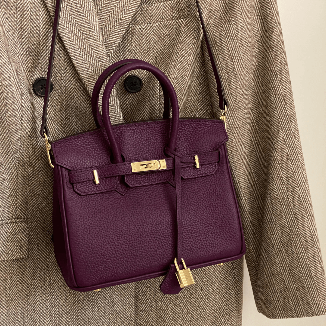 Lila Lederhandtasche mit goldener Hardware, elegant, auf braunem Mantel hängend.