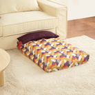 Buntes Hundebett mit geometrischem Muster auf Teppich neben beigem Sofa.