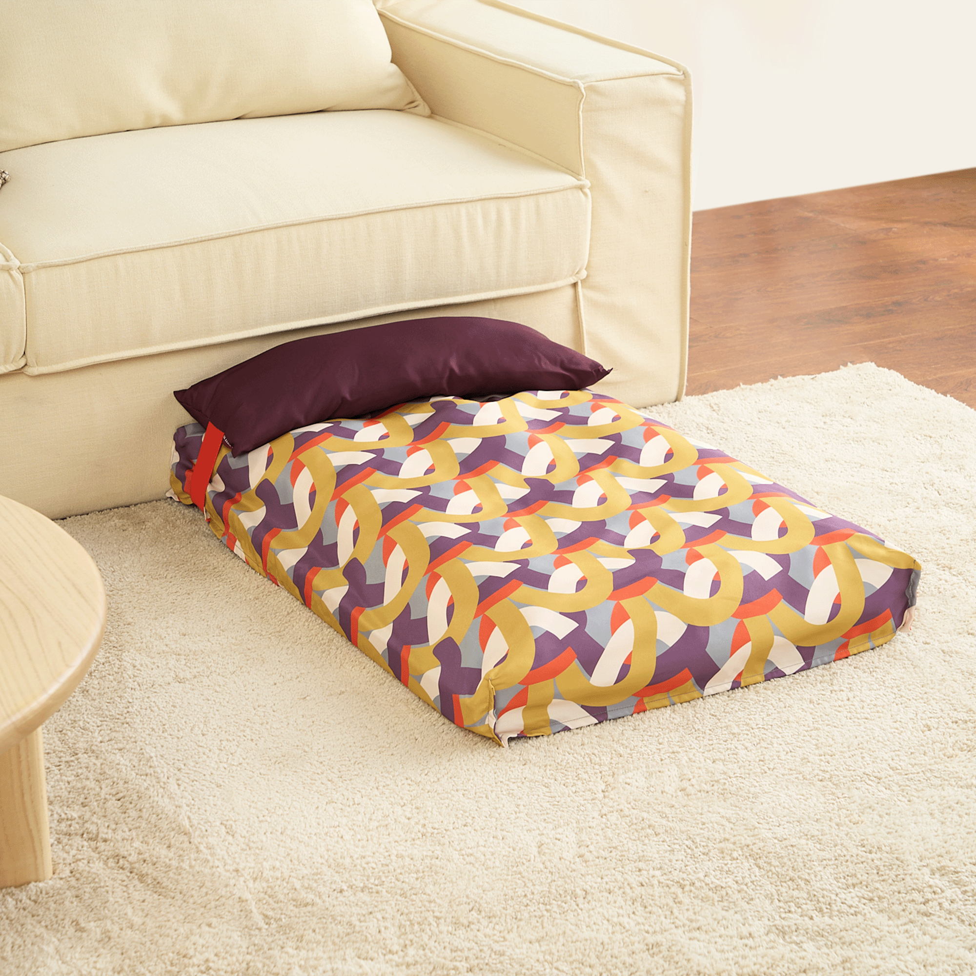 Buntes Hundebett mit geometrischem Muster auf Teppich neben beigem Sofa.