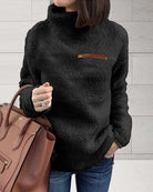 Damenmode: Schwarzer Teddy-Fleece-Pullover mit hohem Kragen und Reißverschlusstasche, kombiniert mit brauner Ledertasche. Wintertrends 2023.